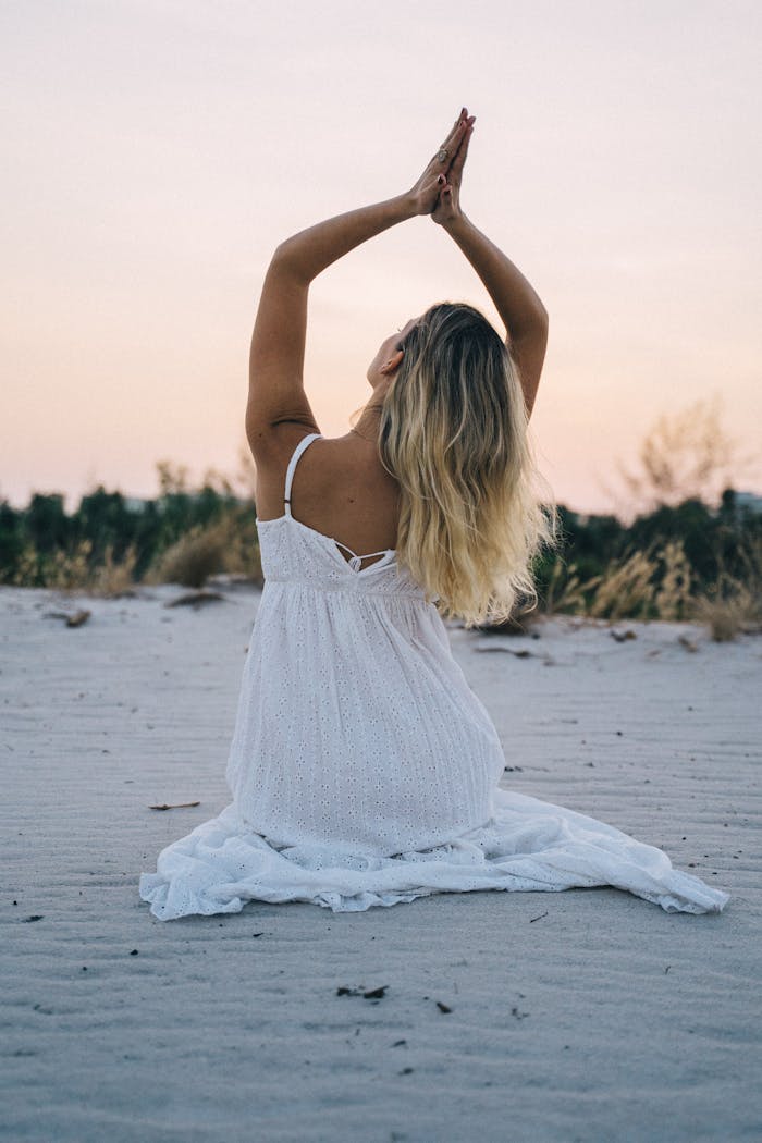Woman in a white dress meditating on a serene beach during sunset, enhancing calmness and mindfulness.